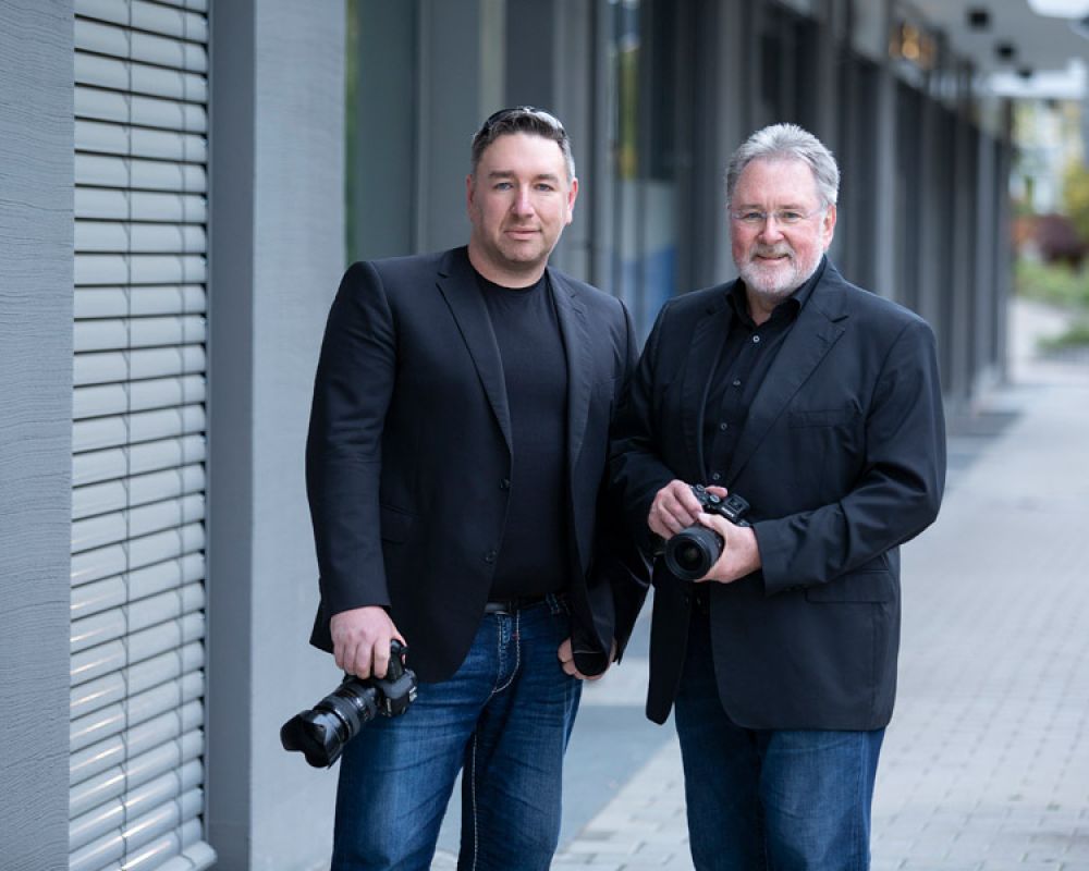 Zwei Fotografen in schwarzer Kleidung mit Kameras, die nebeneinander stehen und in die Kamera schauen.