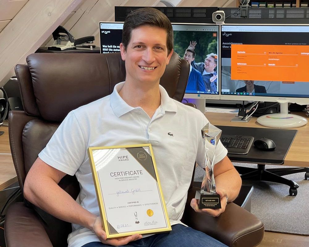 Mann in weißem Polohemd lächelt und hält eine Trophäe sowie ein Zertifikat für den High Performance Award, das seine Auszeichnung und Erfolg zeigt.