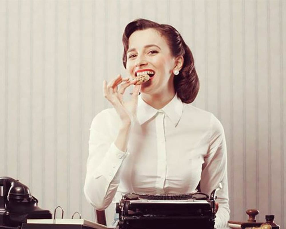 Frau im Vintage-Stil isst einen Keks und sitzt an einer Schreibmaschine, symbolisiert EU-Cookie-Richtlinie oder Telemediengesetz.