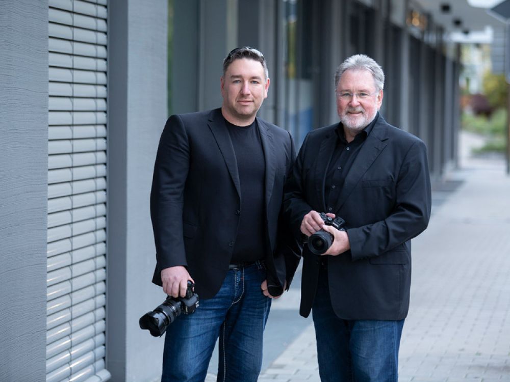 Zwei Fotografen in schwarzer Kleidung mit Kameras, die nebeneinander stehen und in die Kamera schauen.