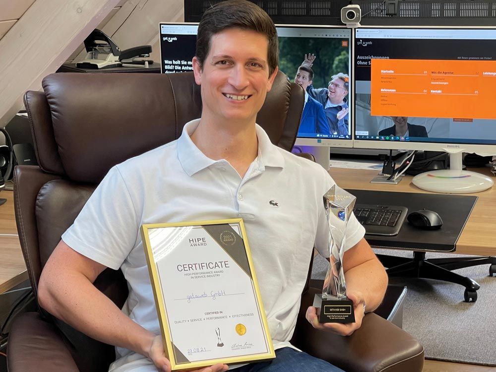 Mann in weißem Polohemd lächelt und hält eine Trophäe sowie ein Zertifikat für den High Performance Award, das seine Auszeichnung und Erfolg zeigt.