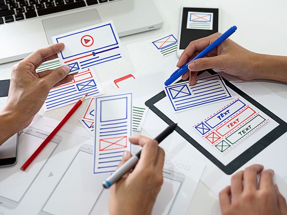 Zwei Personen arbeiten an einem Webdesign-Plan mit handgezeichneten Layouts und Markierungen, verdeutlicht die Bedeutung von strategischem Webdesign in einer Marketingagentur.