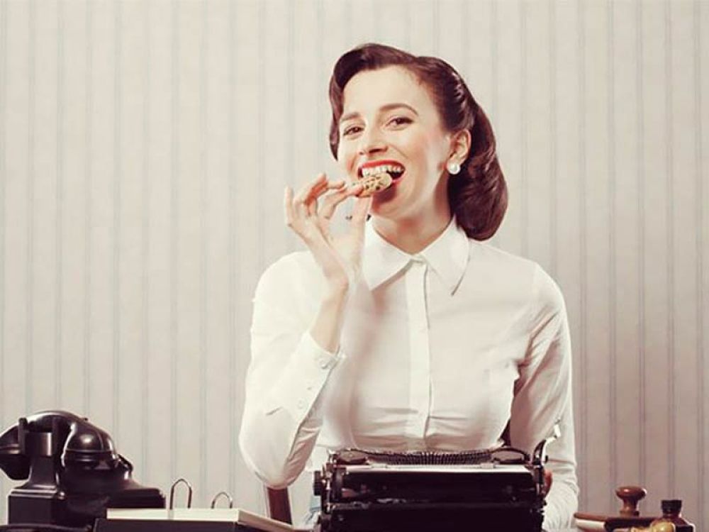 Frau im Vintage-Stil isst einen Keks und sitzt an einer Schreibmaschine, symbolisiert EU-Cookie-Richtlinie oder Telemediengesetz.